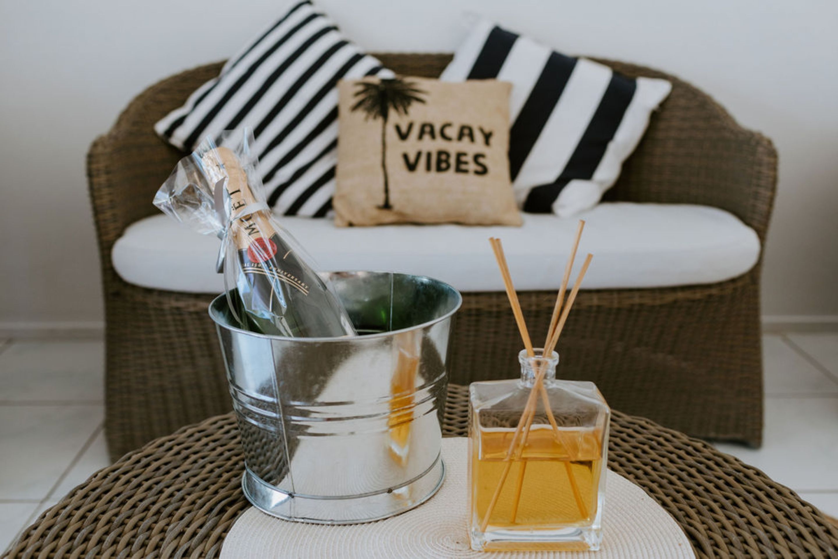 Ein Korbssessel mit gestreiften Kissen und einem Kissen mit der Aufschrift 'VACAY VIBES', ein Metallkübel mit einer Flasche und eine Glasflasche mit Schilfrohrchen.