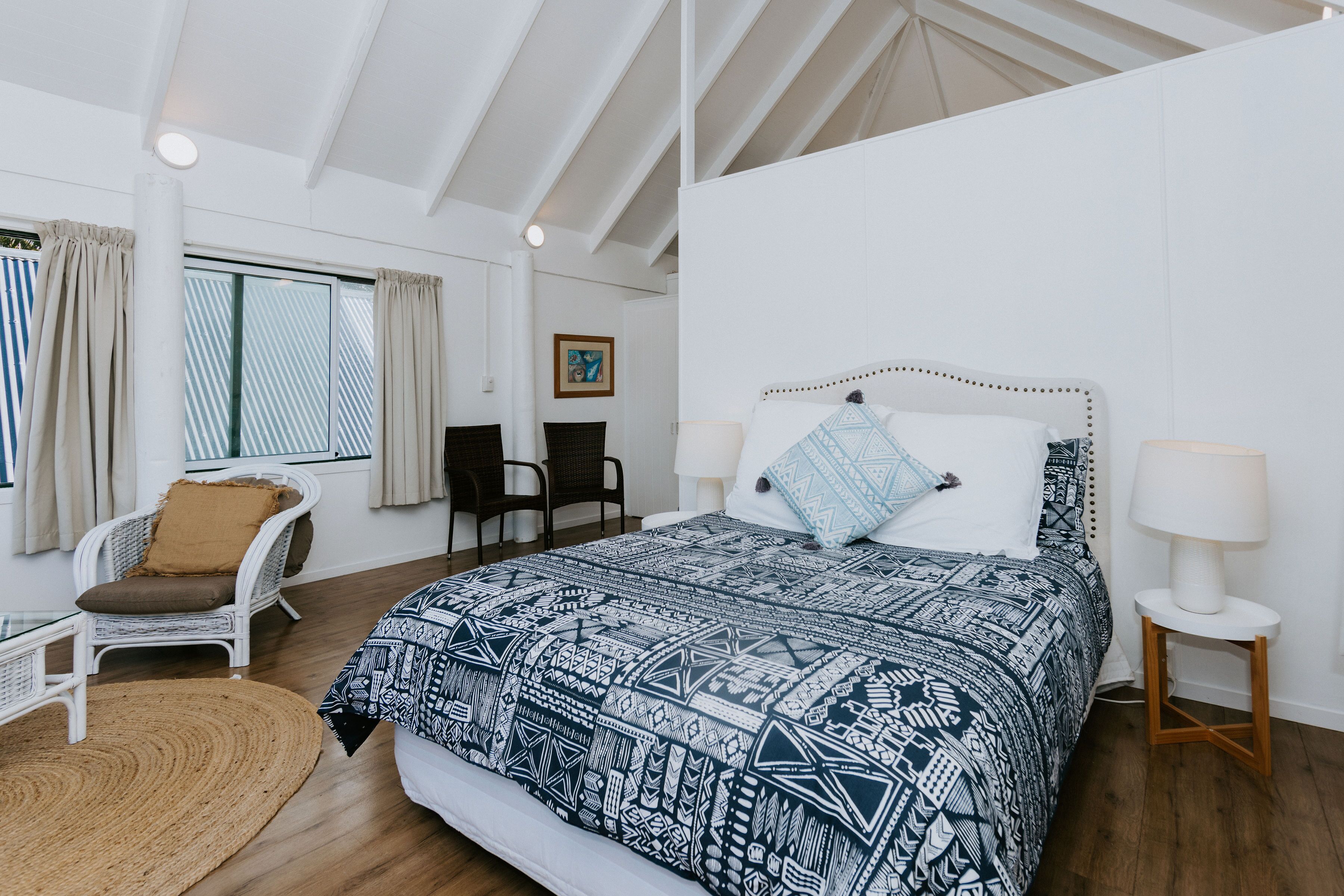 Ein Schlafzimmer mit einem großen Bett, einem Korbstuhl, einem kleinen Tisch und einem Fenster mit Vorhängen.