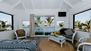 A room with a bed, two white wicker chairs, a white sofa, a small table with a vase of flowers, and large windows with a view of palm trees and the ocean. - The Passage Villas (Rarotonga)
