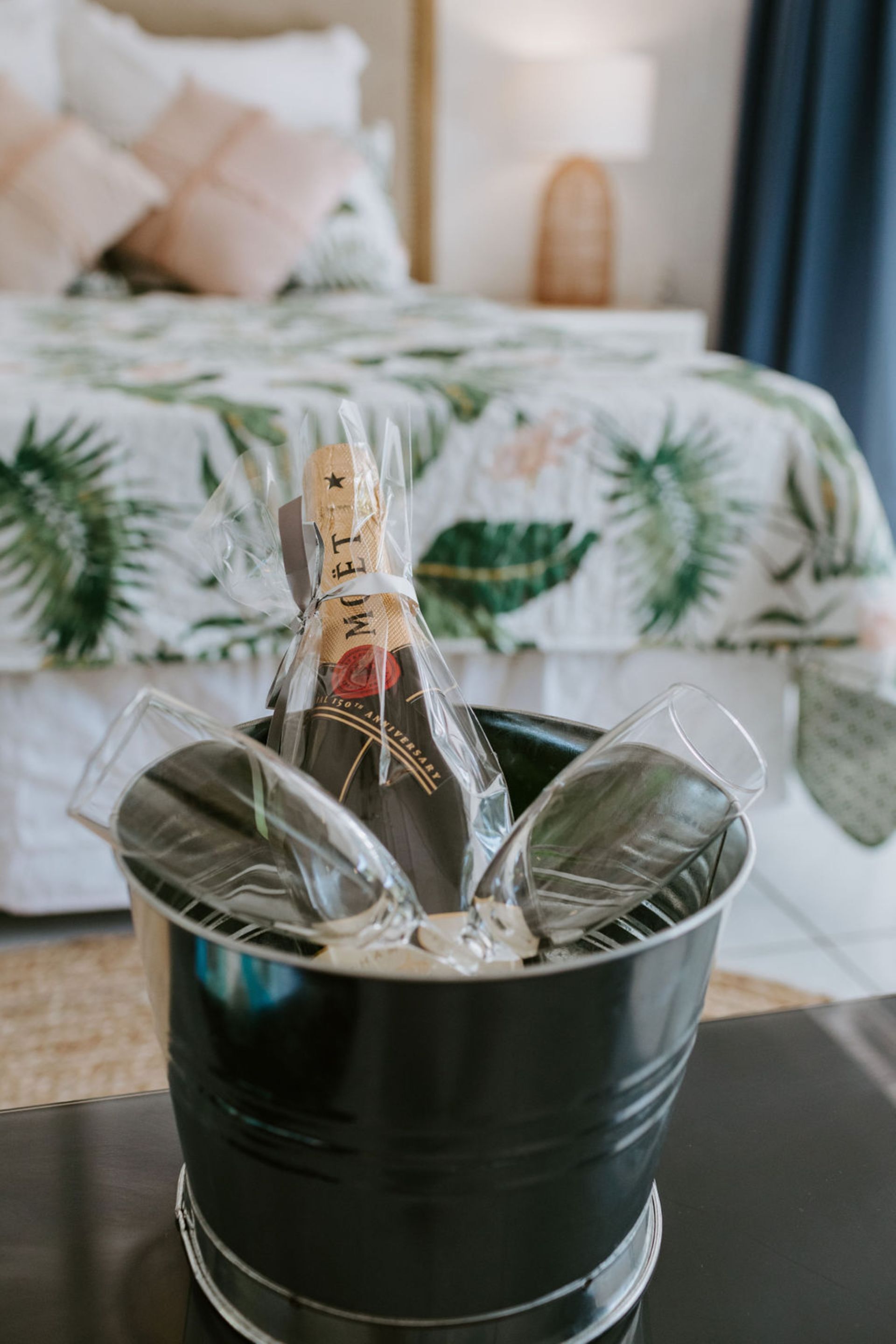 A bed with a floral bedspread and pillows, a black bucket with a bottle of champagne and glasses, and a lamp in the background.