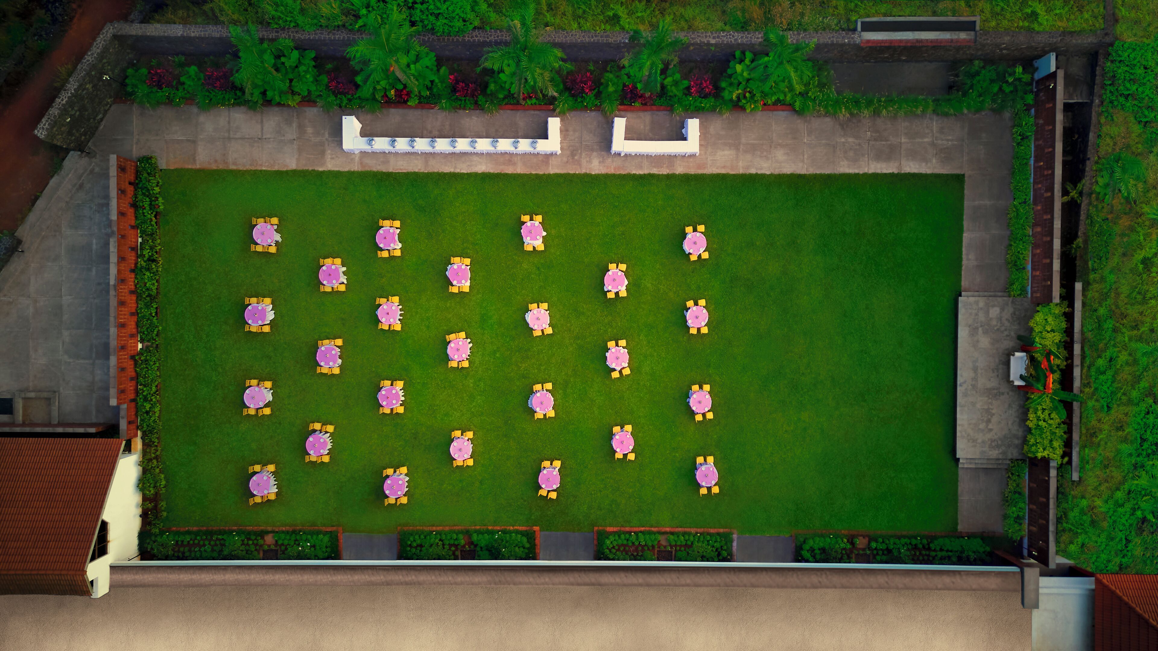 An aerial view of a courtyard with multiple tables and chairs arranged in a grid pattern, surrounded by a low stone wall and lush greenery.