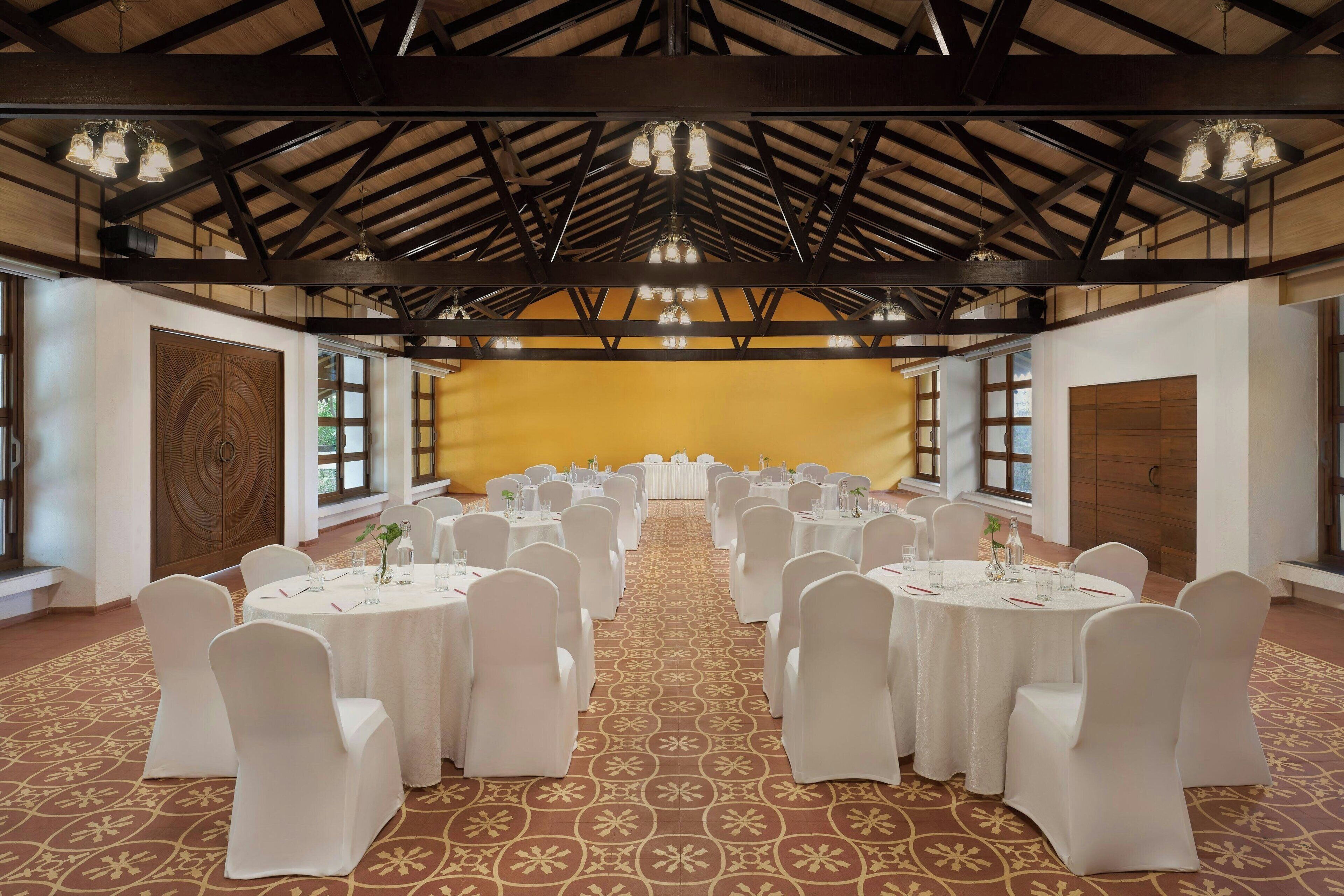 Une salle de banquet avec des tables rondes et des chaises blanches, de grandes fenêtres et un plafond haut avec des poutres apparentes.