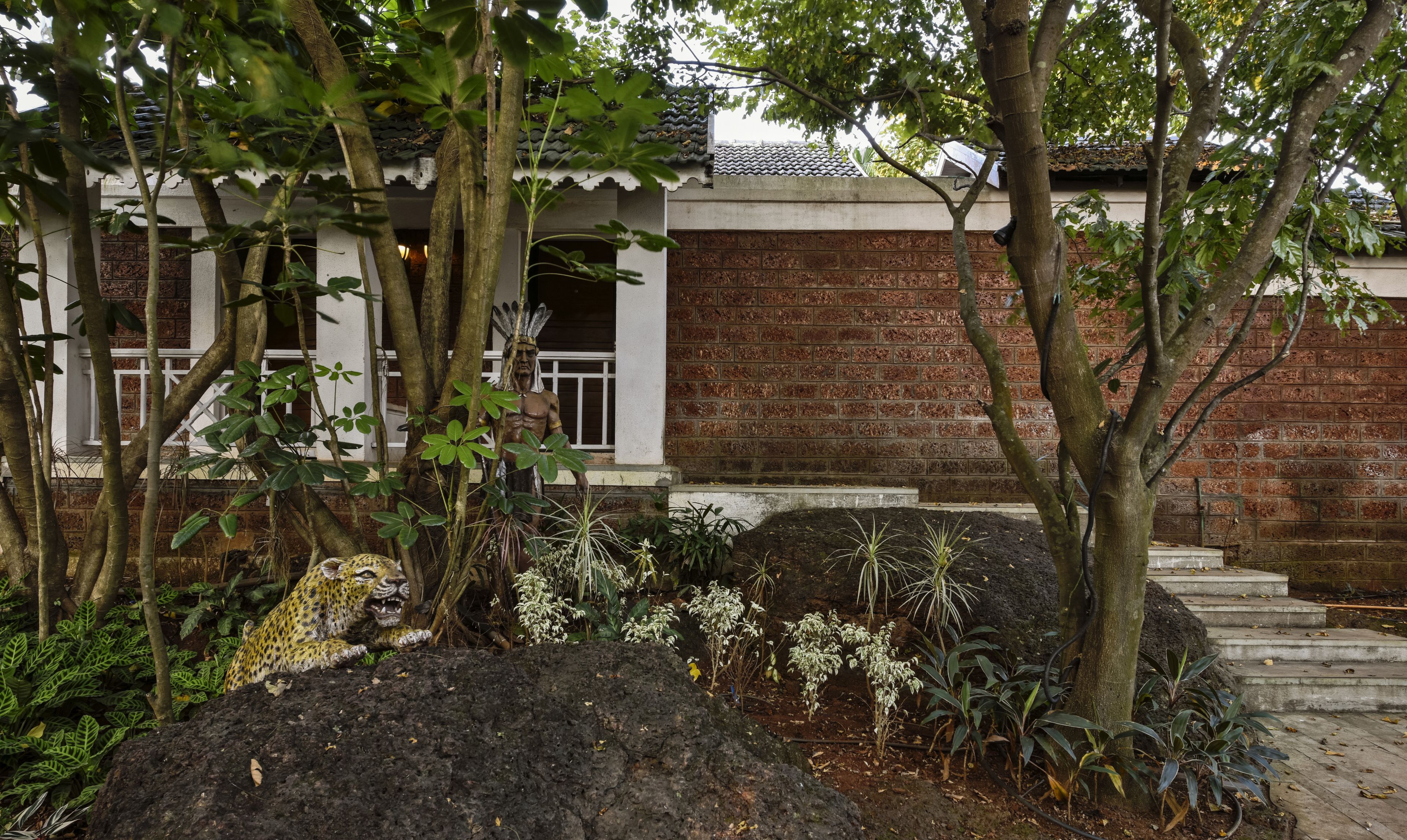 Un edificio tradicional con fachada de ladrillo y tejado de tejas, rodeado de abundante vegetación y un jardín ajardinado con rocas grandes y diversas plantas.