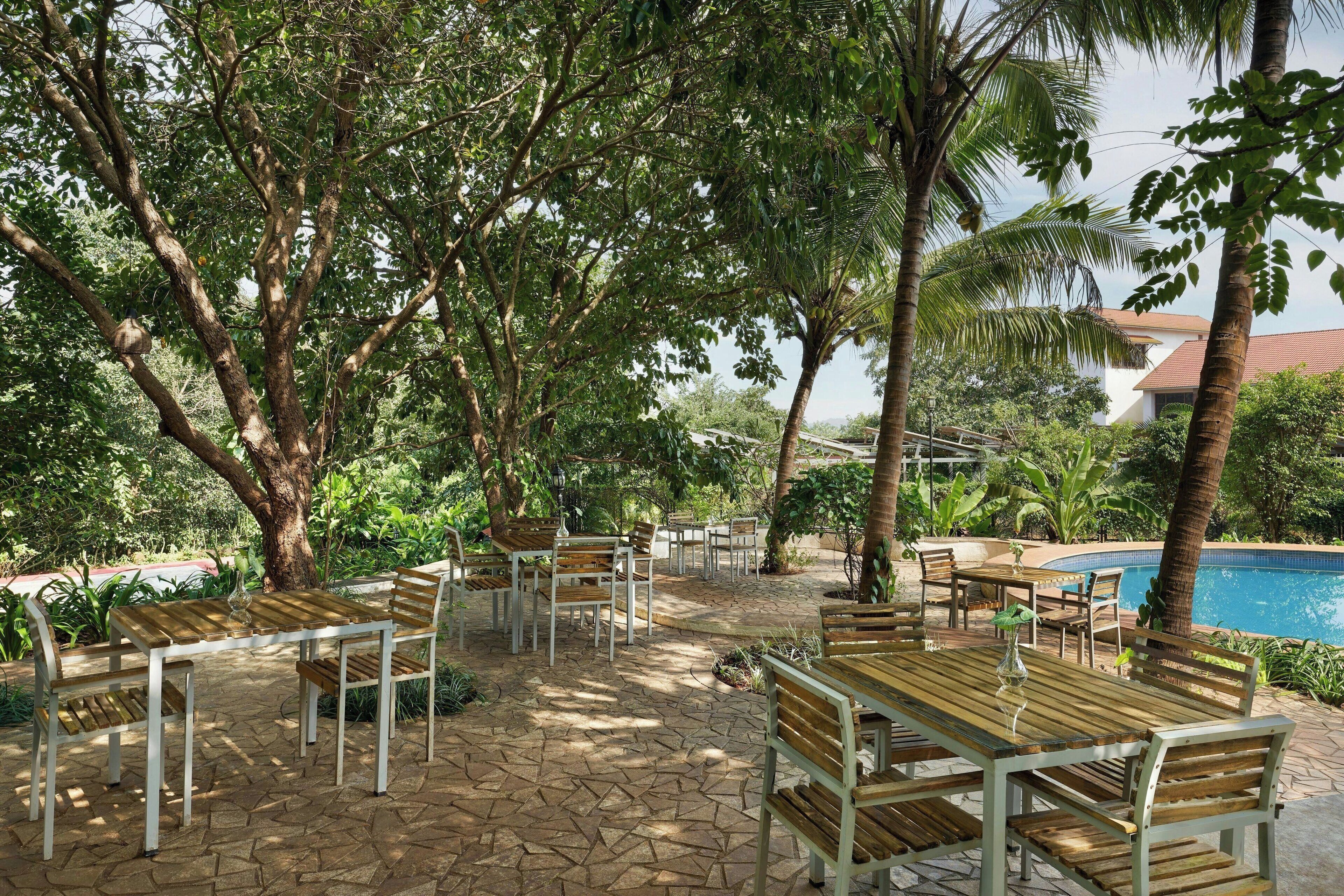 An outdoor patio with wooden tables and chairs, surrounded by lush greenery and a pool in the background.