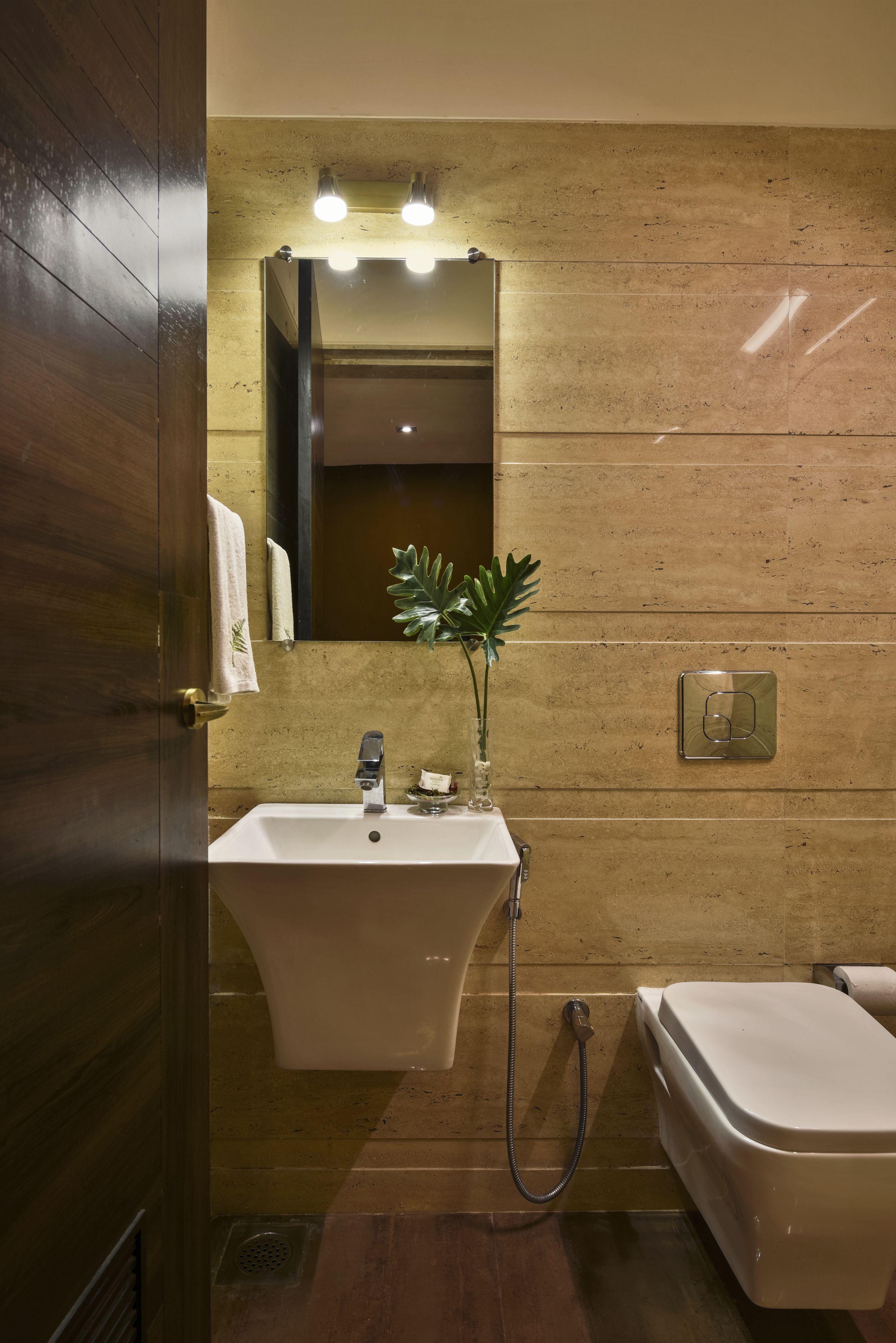 A bathroom with a wooden door, a sink with a mirror above it, a plant, and a toilet.