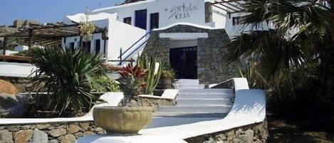 Un edificio blanco con base de piedra y puerta azul, rodeado de plantas tropicales y un cielo despejado.