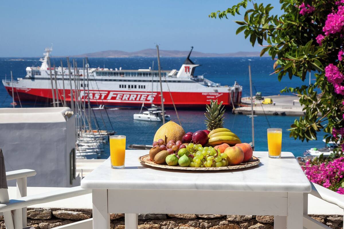 A table with fruits and a glass of orange juice, overlooking a marina with boats and a ferry.