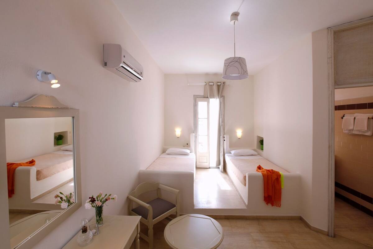 A hotel room with two single beds, a sitting area with a chair and table, a mirror, and a hanging light fixture.