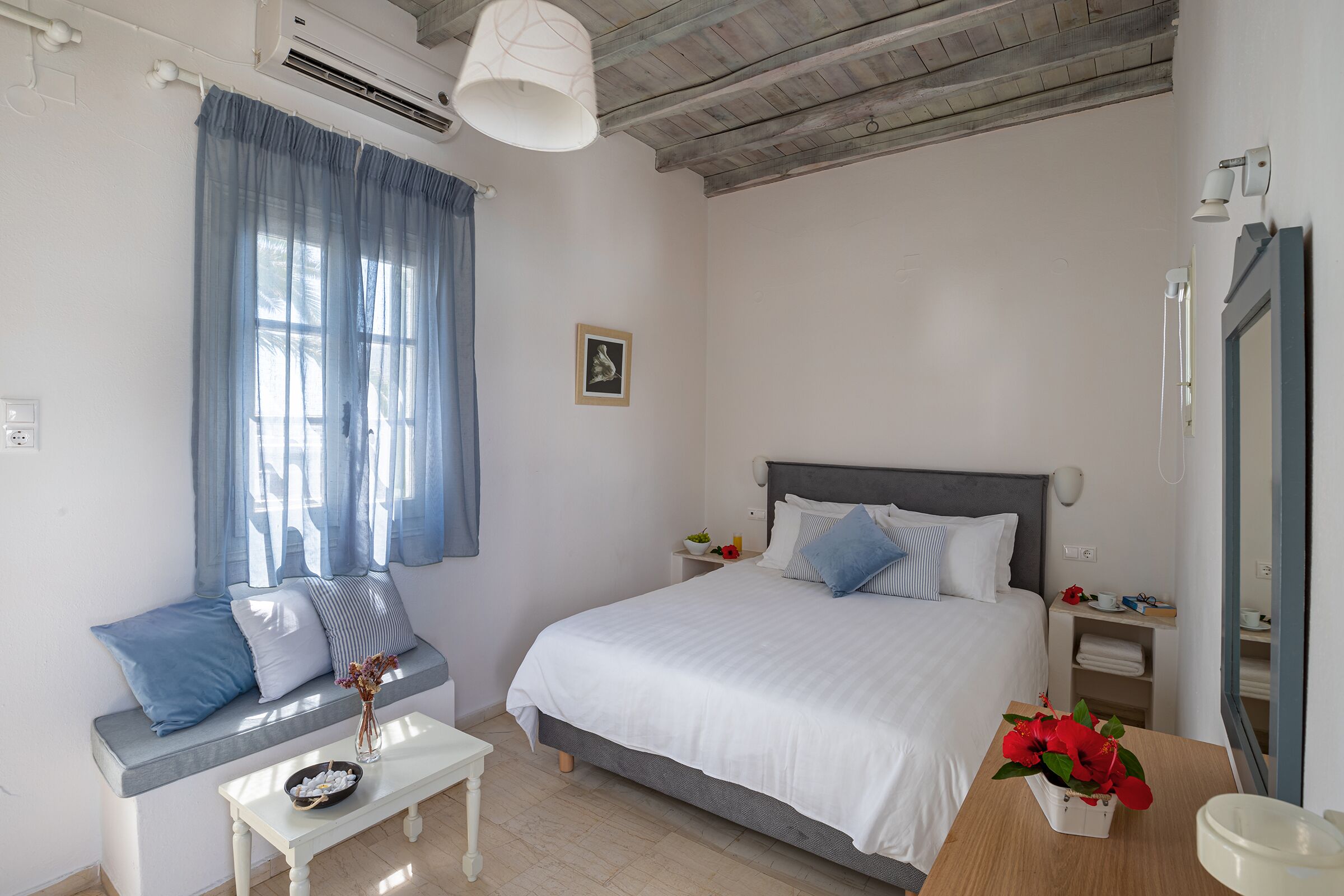 A bedroom with a bed, a nightstand, a bedside table, a window with curtains, and a wall-mounted air conditioner.