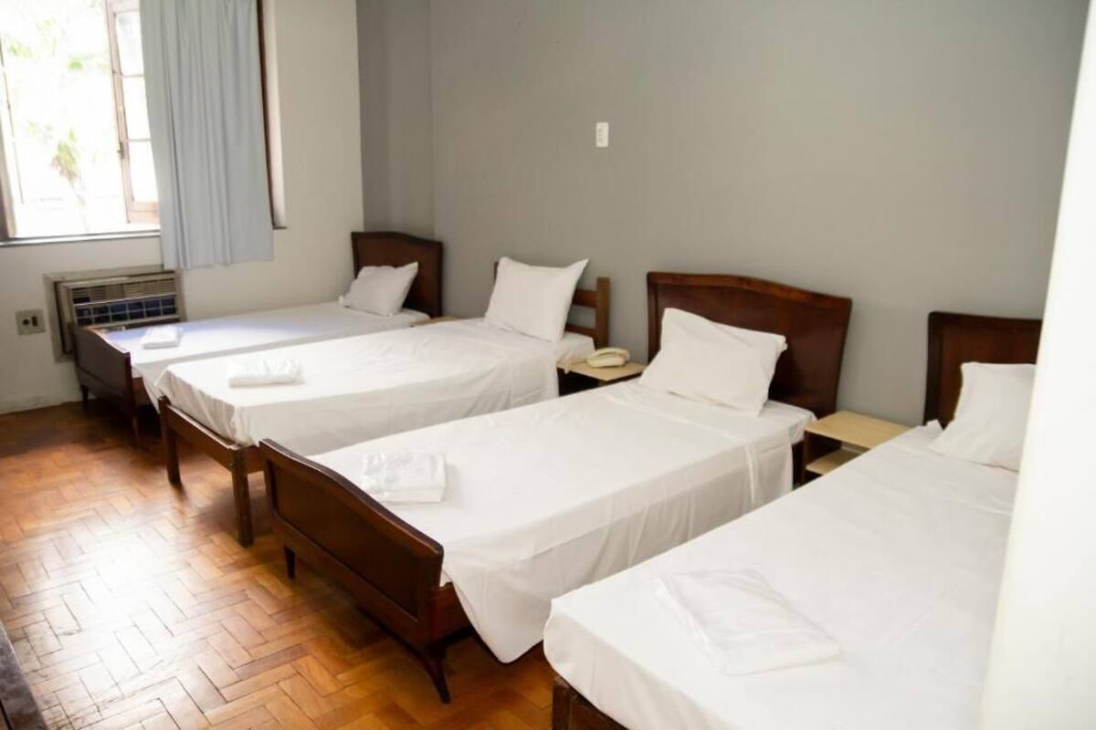 A hotel room with three single beds, wooden headboards, white linens, a small bedside table with a lamp, and a window with light curtains.