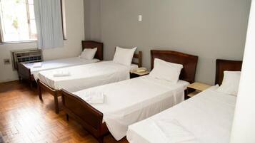 Habitación de hotel con tres camas individuales, cabeceros de madera, ropa de cama blanca, una mesita de noche pequeña con lámpara y una ventana con cortinas claras.