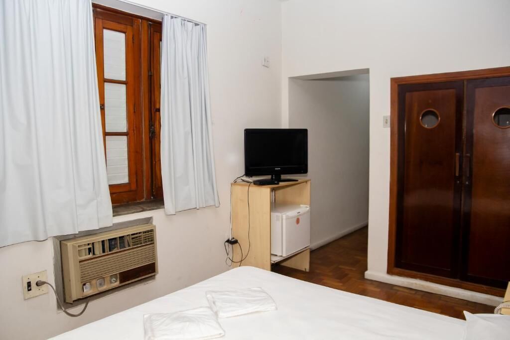 Een kleine, eenvoudige kamer met een bed, een televisie op een houten standaard, een kleine koelkast en een kledingkast.