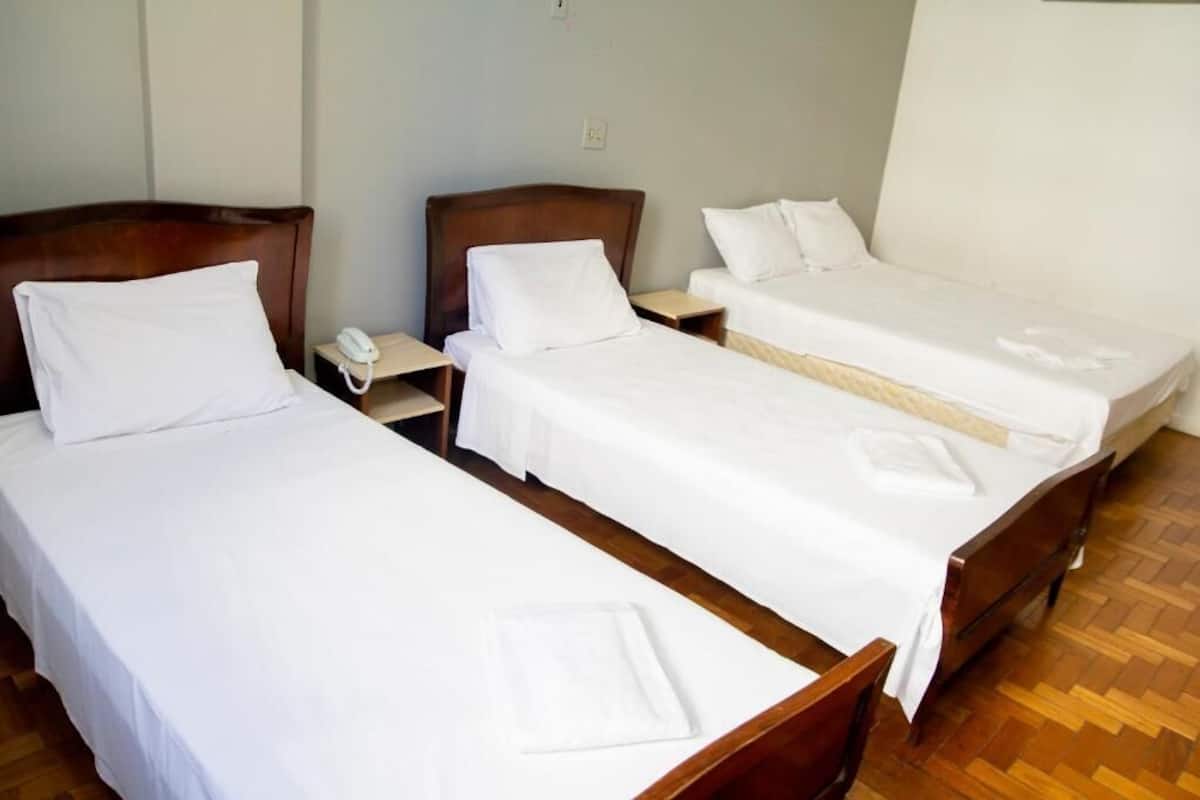 A hotel room with three single beds, wooden headboards, white bedding, and a wooden floor.