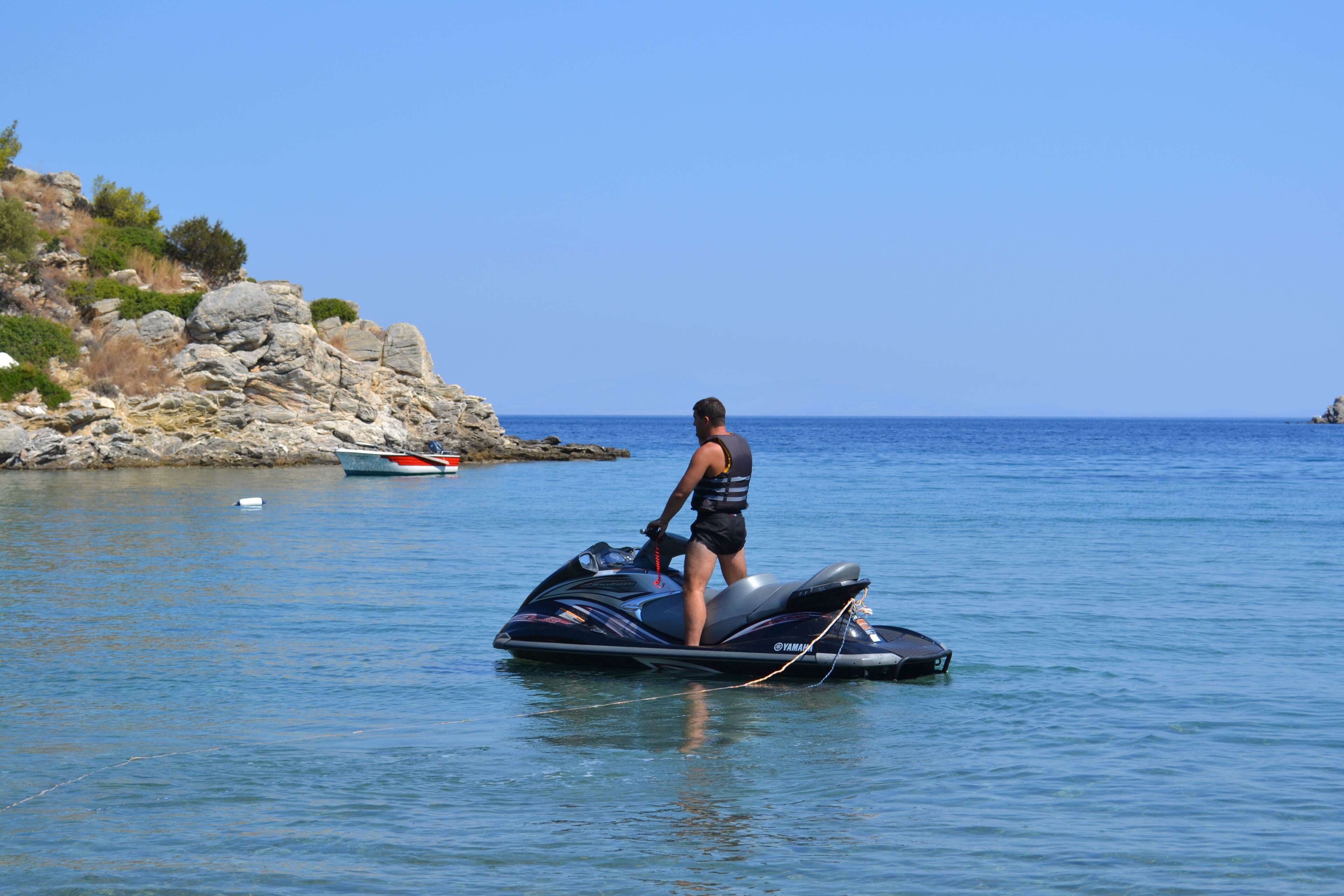 Una persona en un jet ski sobre aguas tranquilas, con una costa rocosa y un cielo despejado al fondo.