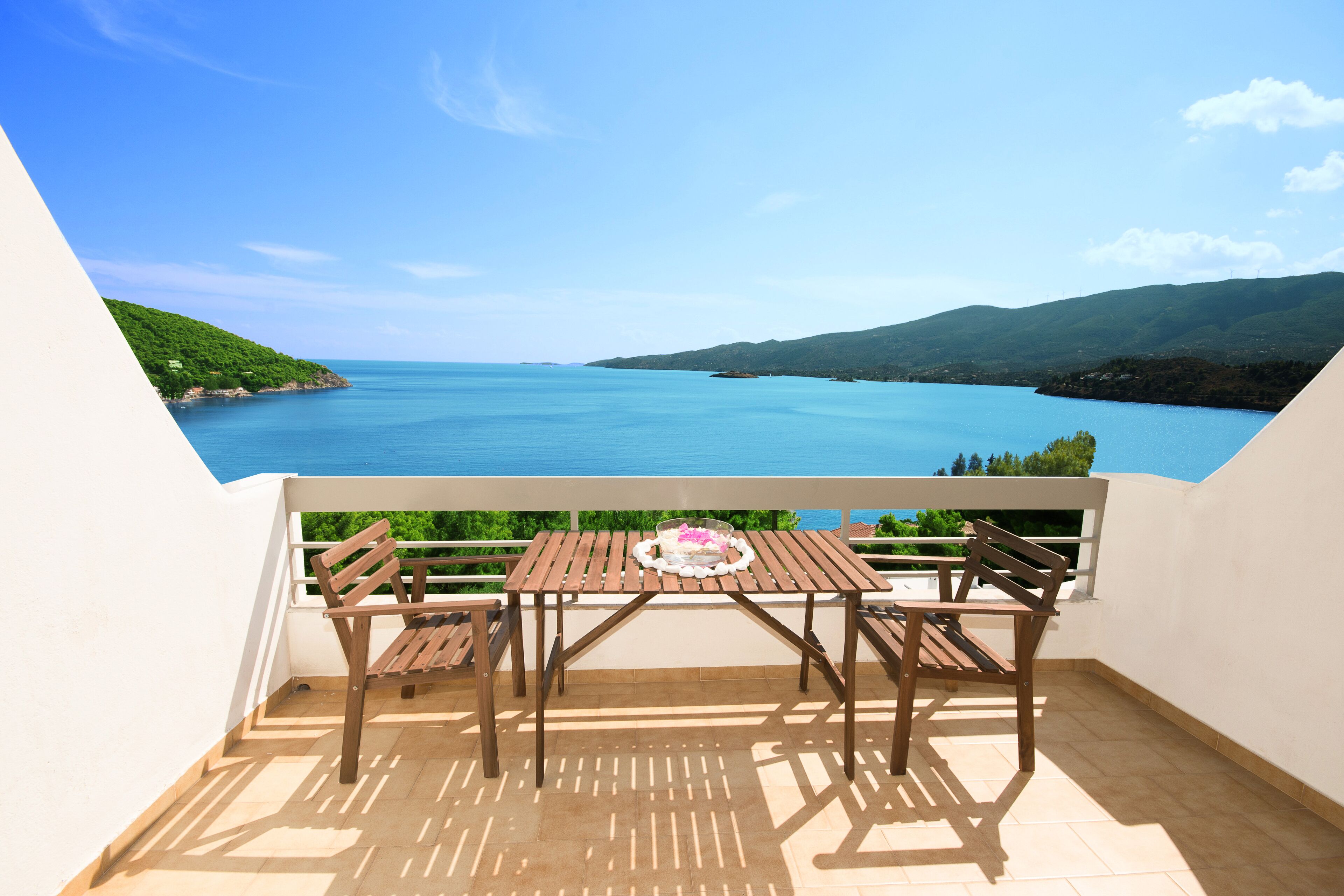 A balcony with a wooden table and chairs, overlooking a large body of water and green hills.