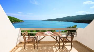 A balcony with a wooden table and chairs, overlooking a large body of water and green hills.