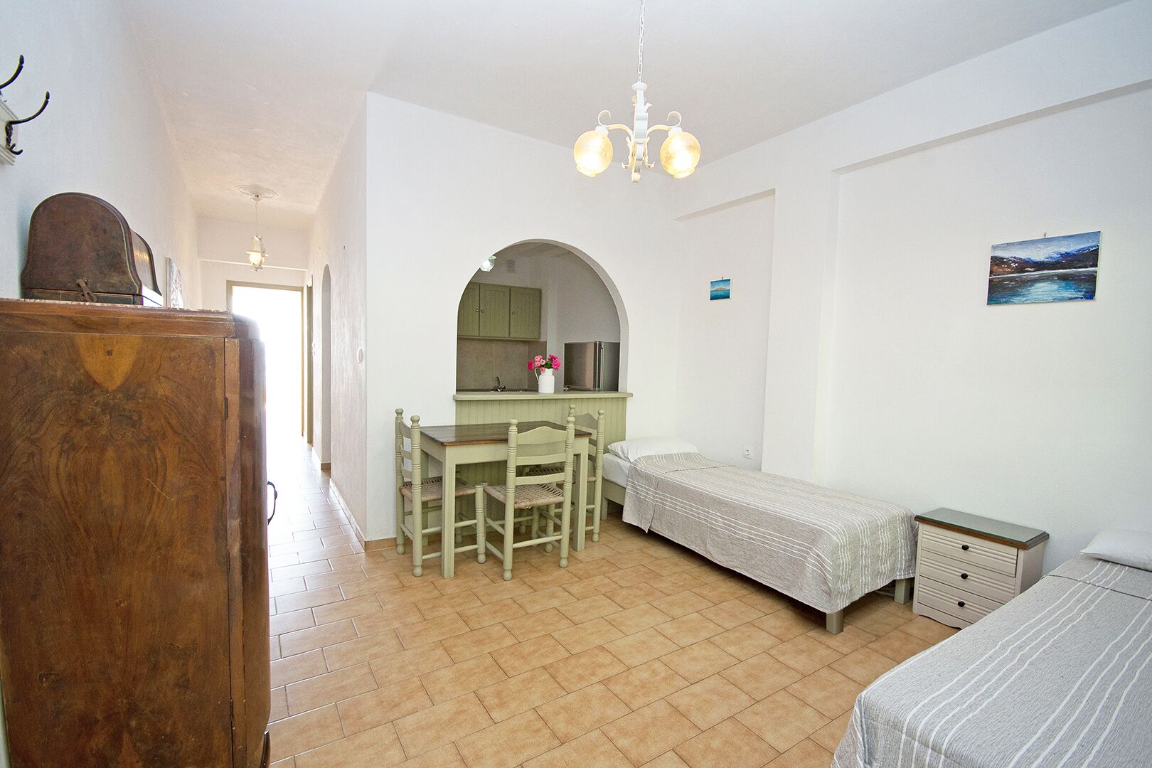 Habitación compacta con armario de madera, una pequeña mesa de comedor con sillas, una cama individual y un rincón con kitchenette.