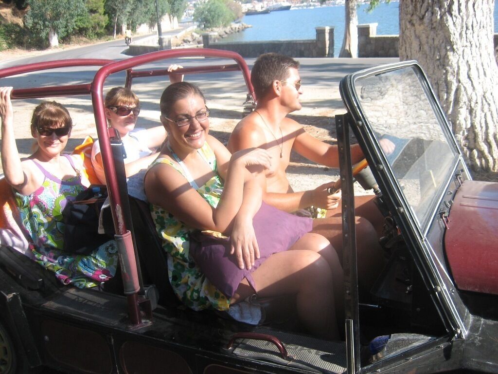A vintage car with passengers, parked near a waterfront.