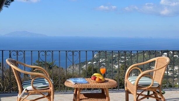 A balcony with wicker chairs and a small table, overlooking a scenic view of the sea and distant land. - Alle Ginestre B&B (Anacapri)