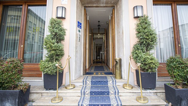 A hotel entrance with a blue carpet, two potted trees, and a sign indicating a room rate. - Hotel PRime già Hotel Piccolo Ritz (Pistoia)