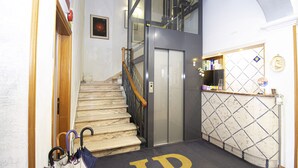 A hotel lobby with a marble staircase, an elevator, and a reception desk with a decorative mirror. - Hotel PRime già Hotel Piccolo Ritz (Pistoia)
