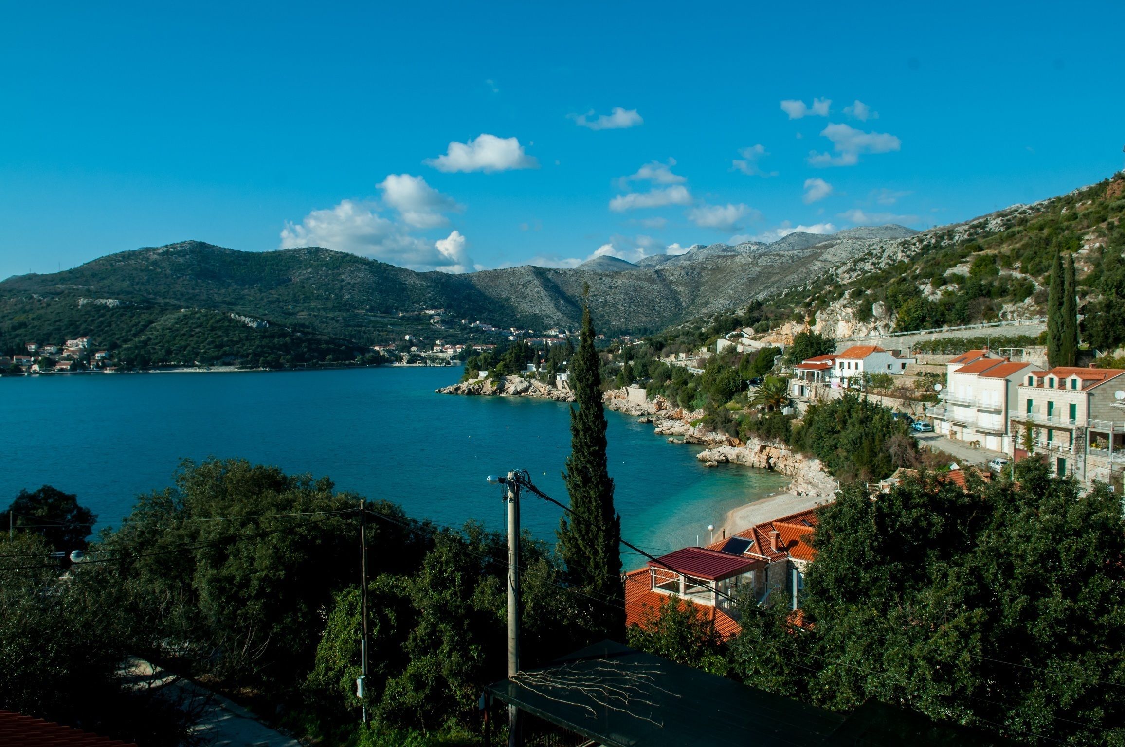 A coastal town with buildings along the shore, a clear blue sea, and mountains in the background.