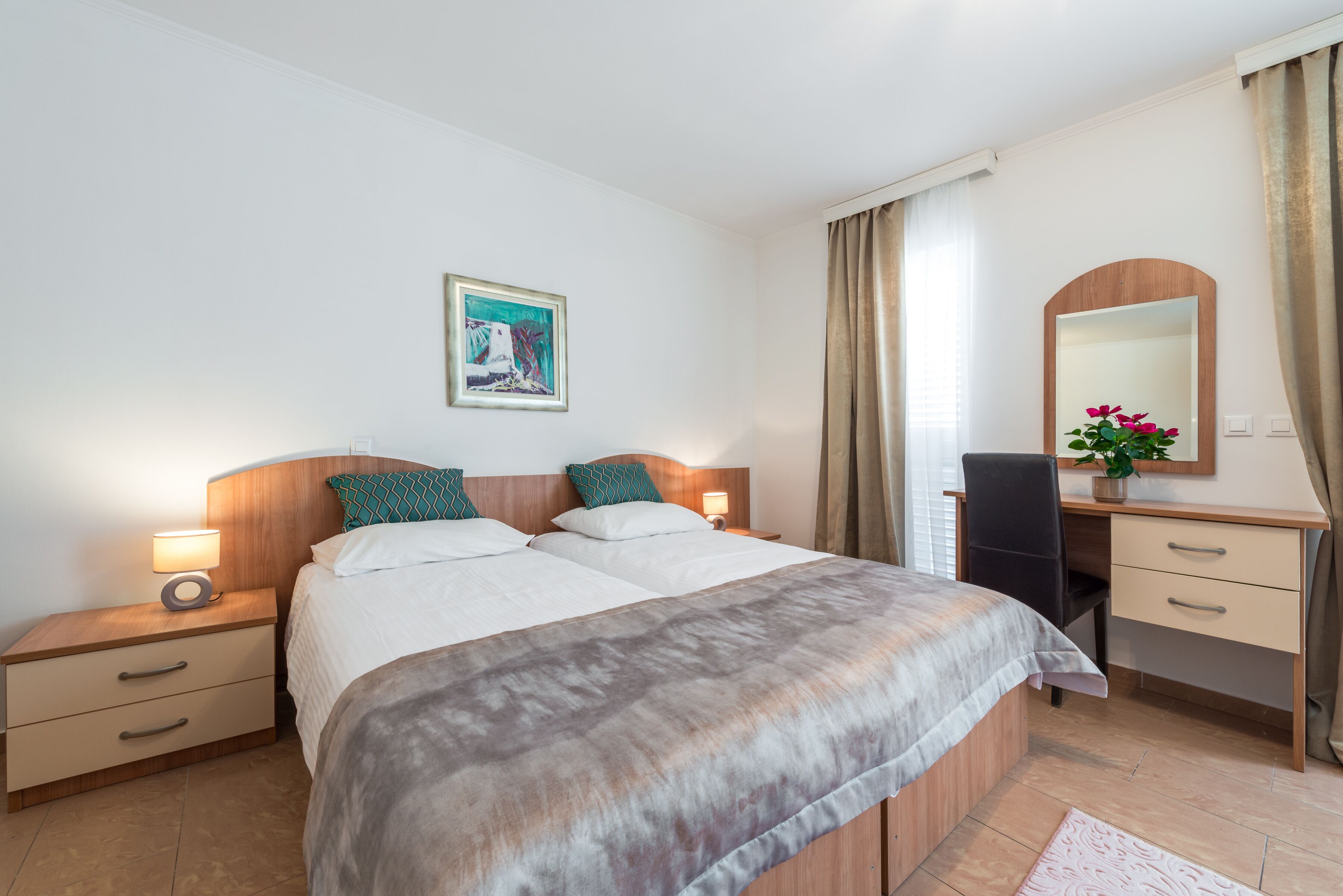 A hotel room with a bed, bedside tables, a desk, a chair, a mirror, and a painting on the wall.