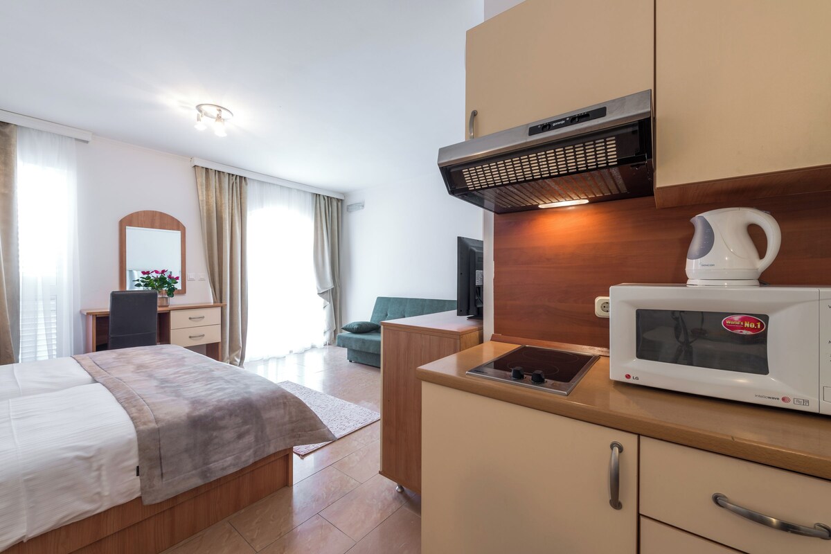 A modern hotel room with a bed, a kitchenette with a microwave, a sink, and a wooden cabinet. There is a desk with a chair and a TV in the corner.