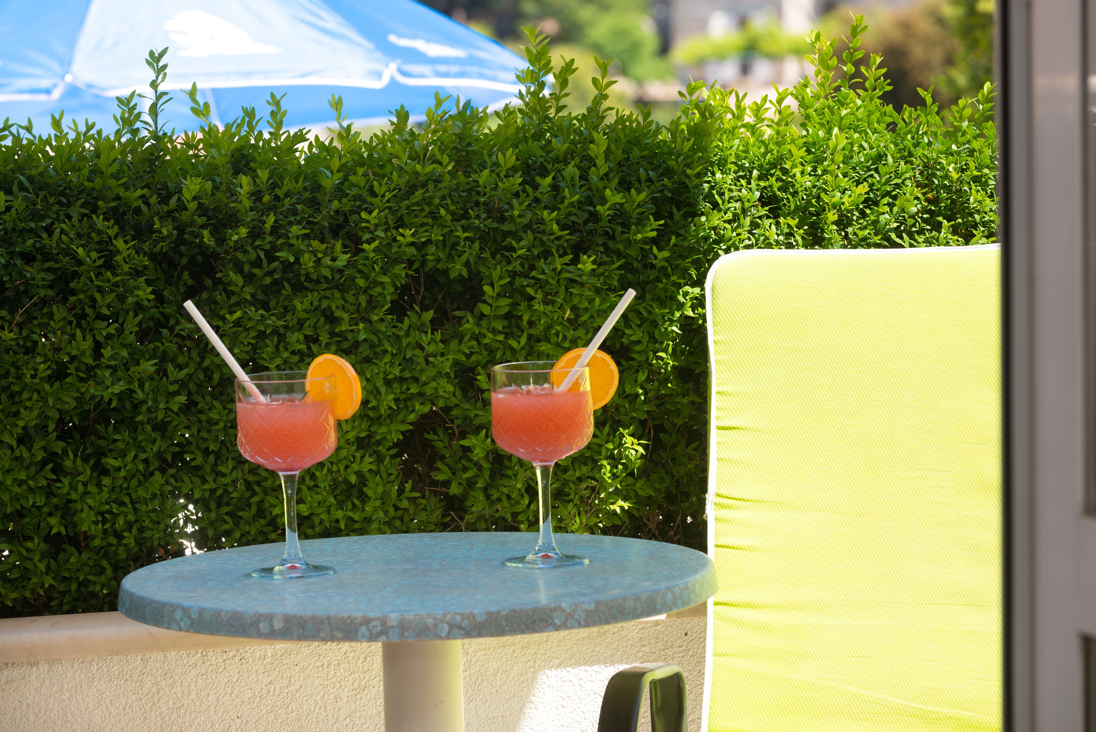 Two cocktails on a round table with a green hedge in the background.