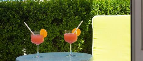 Two cocktails on a round table with a green hedge in the background.