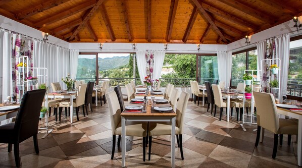 A dining area with tables set for four, large windows offering a view of the outdoors, and a wooden ceiling.