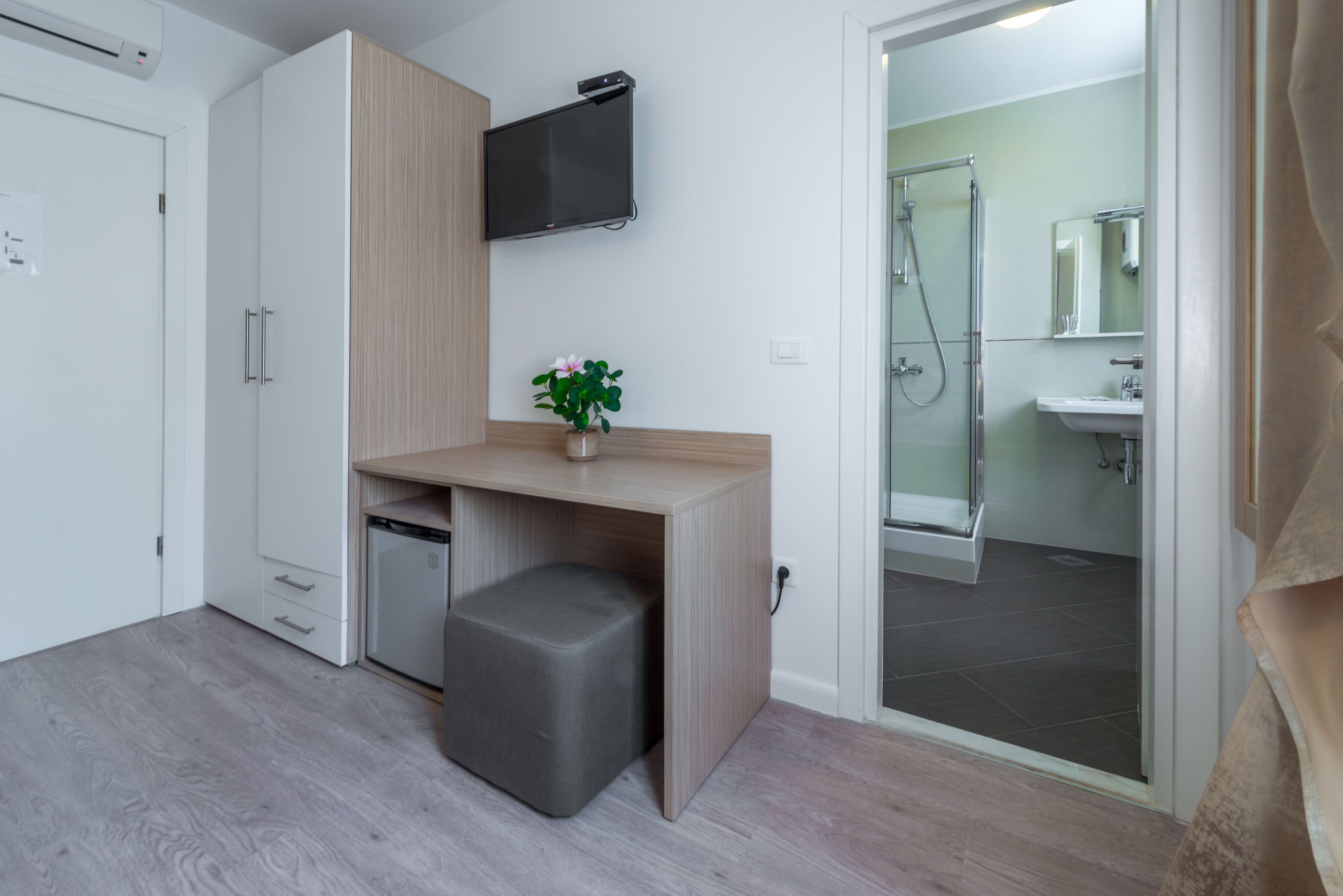 A compact room with a kitchenette, a small seating area, a TV mounted on the wall, and a shower area visible through an open door.