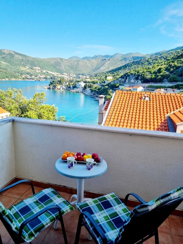 A balcony with a table set with fruit and two chairs, overlooking a lake and mountains.