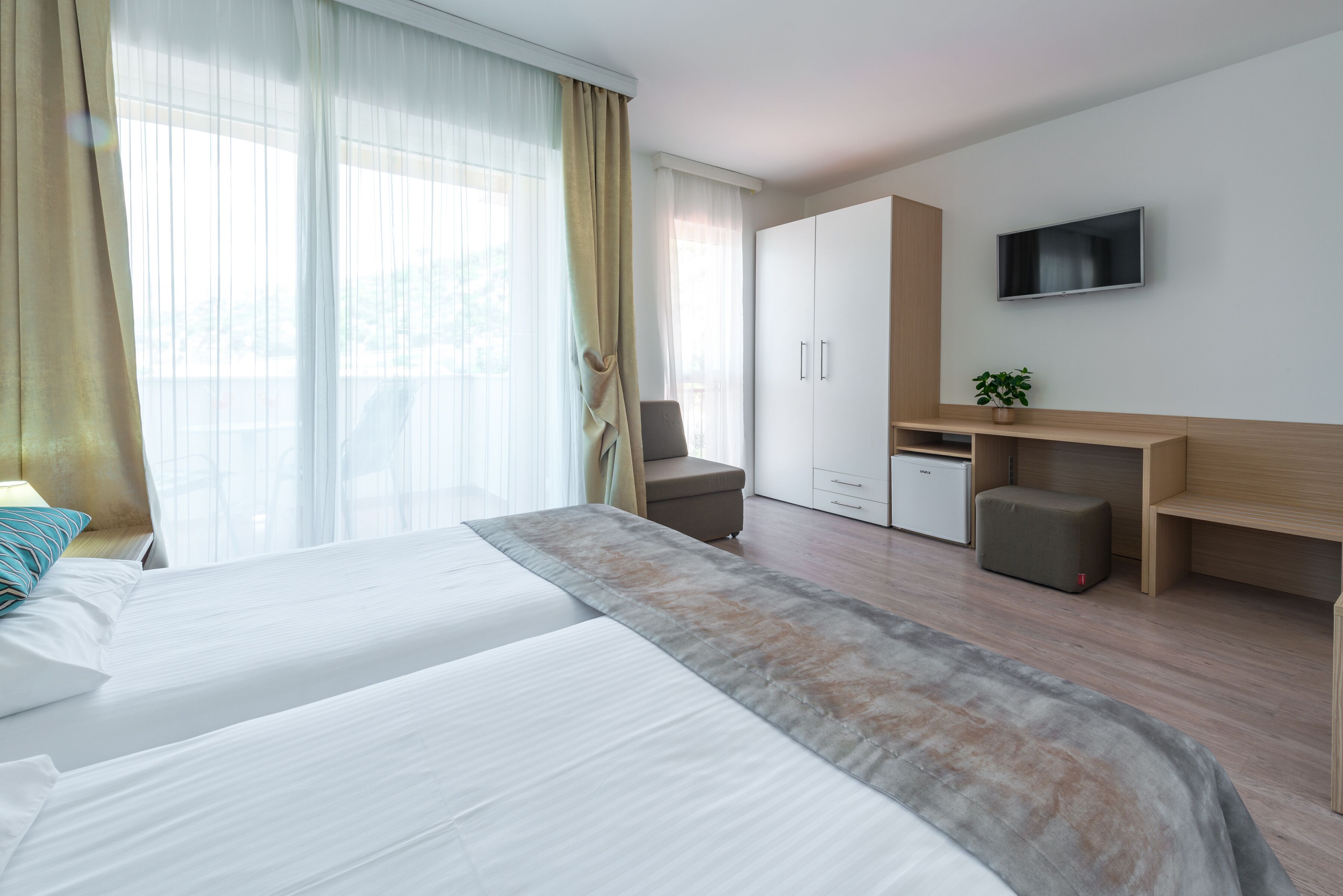 A hotel room with a large bed, a desk, a chair, a TV, and a small plant.
