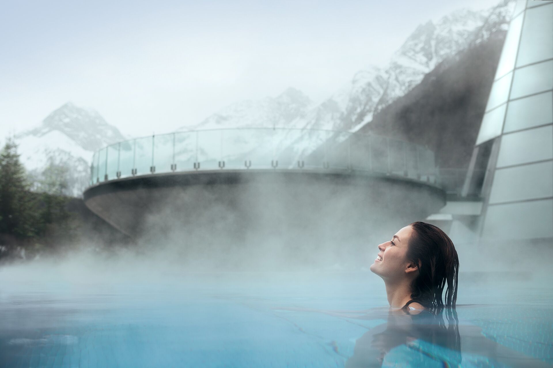 Uma pessoa em uma fonte termal com vapor subindo, cercada por um edifício moderno e montanhas nevadas ao fundo.