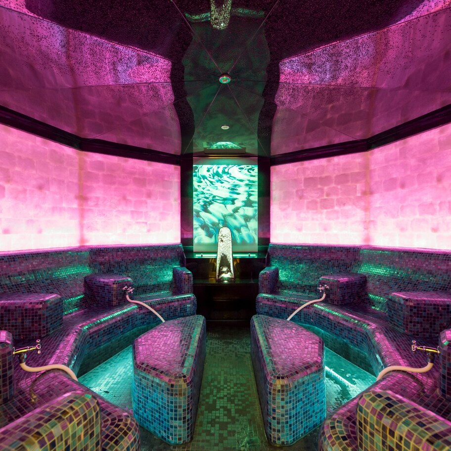 A spa room with mosaic-tiled walls and benches, a central water feature, and a ceiling with a geometric design.
