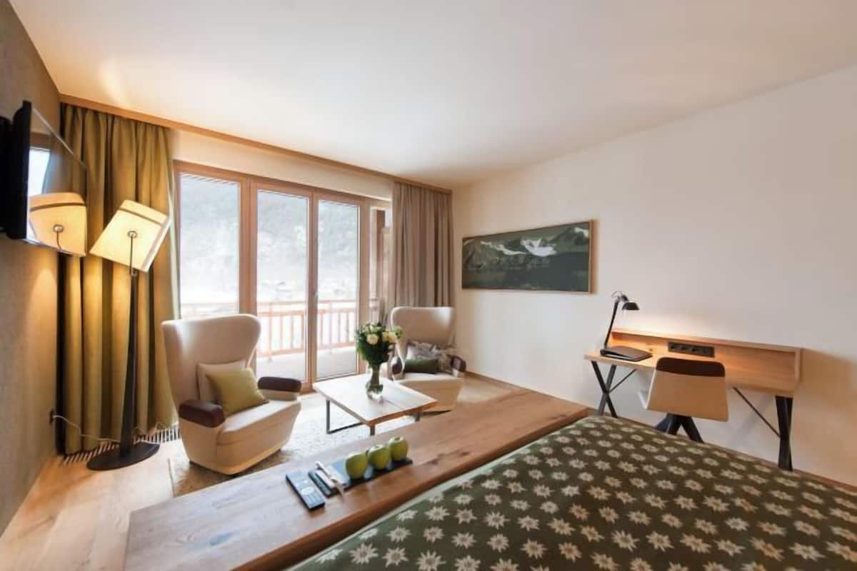 A hotel room with a bed, two chairs, a desk, a TV, and a window with curtains.