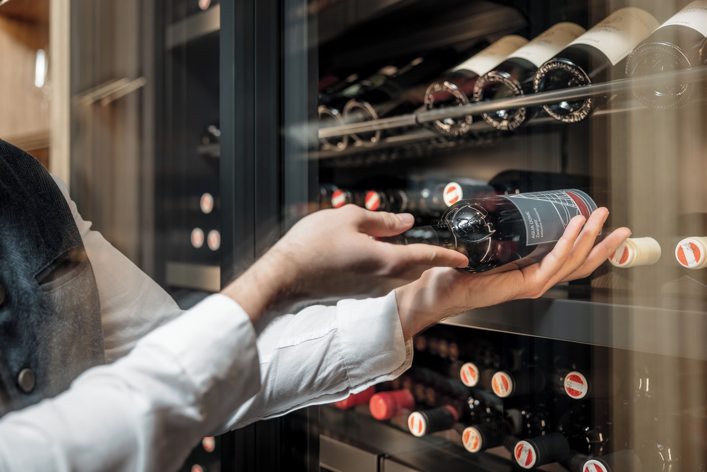 Una persona sosteniendo una botella de vino frente a una estantería para vinos.