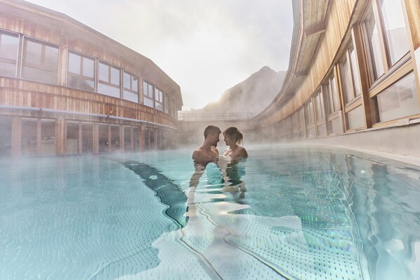 A couple in a clear indoor pool with large windows and a wooden building in the background.