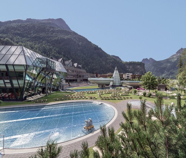 A modern hotel with a glass facade, a pool area, and a mountain backdrop.