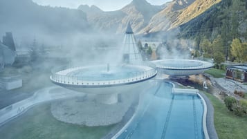 Un spa termal con una piscina circular y una fuente central, rodeado de montañas y niebla.
