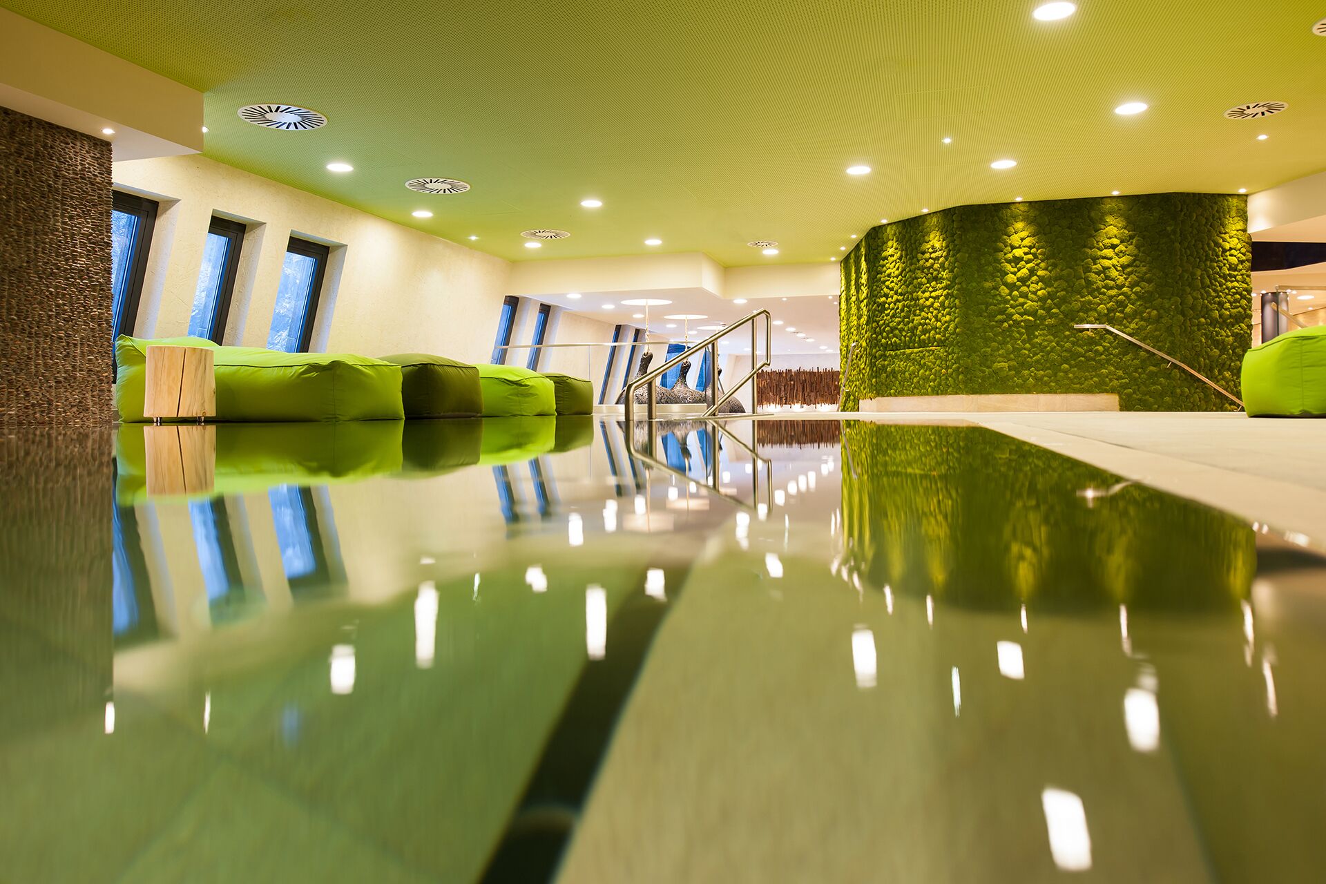 Un área de piscina interior moderna con piso reflectante, asientos verdes y una pared de piedra con diseño de mosaico verde.