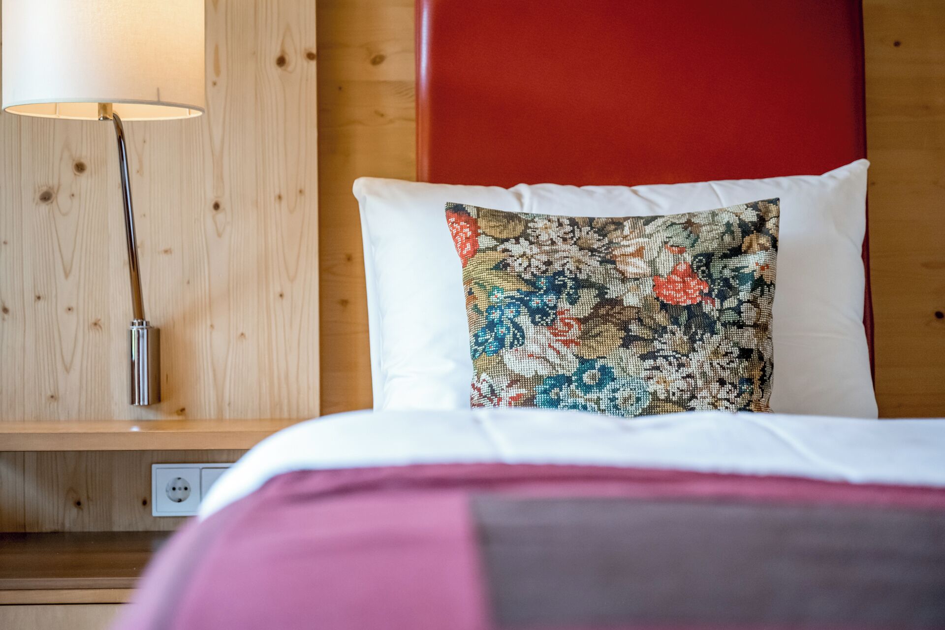 Une chambre d’hôtel avec une tête de lit en bois, un coussin à motifs floraux, une lampe de chevet et un lit recouvert d’une couverture rose et grise.