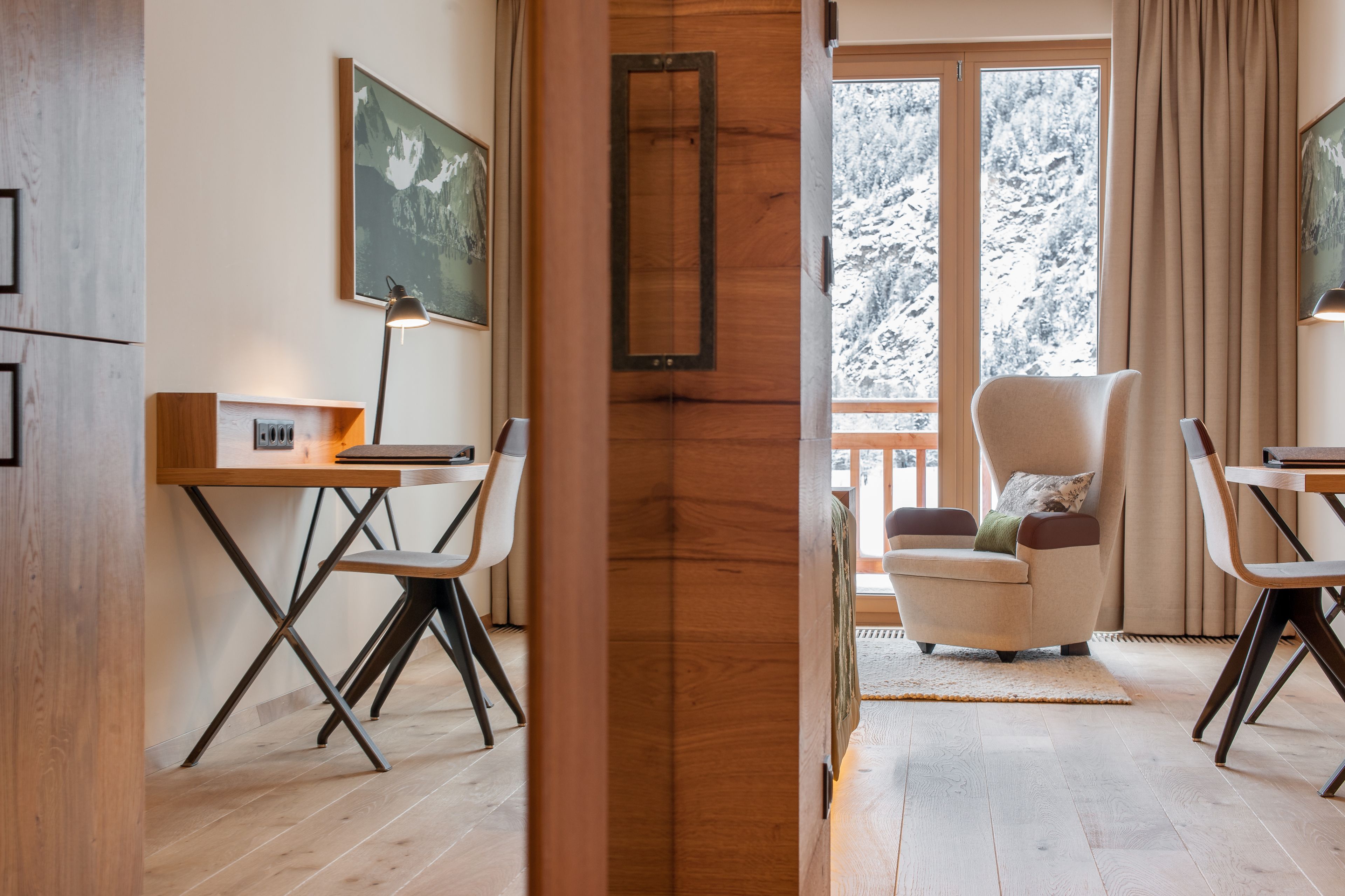 Ein modernes Wohnzimmer mit Holzboden, einem Schreibtisch mit Lampe, einem gemütlichen Sessel und einem großen Fenster mit Blick auf schneebedeckte Bäume.