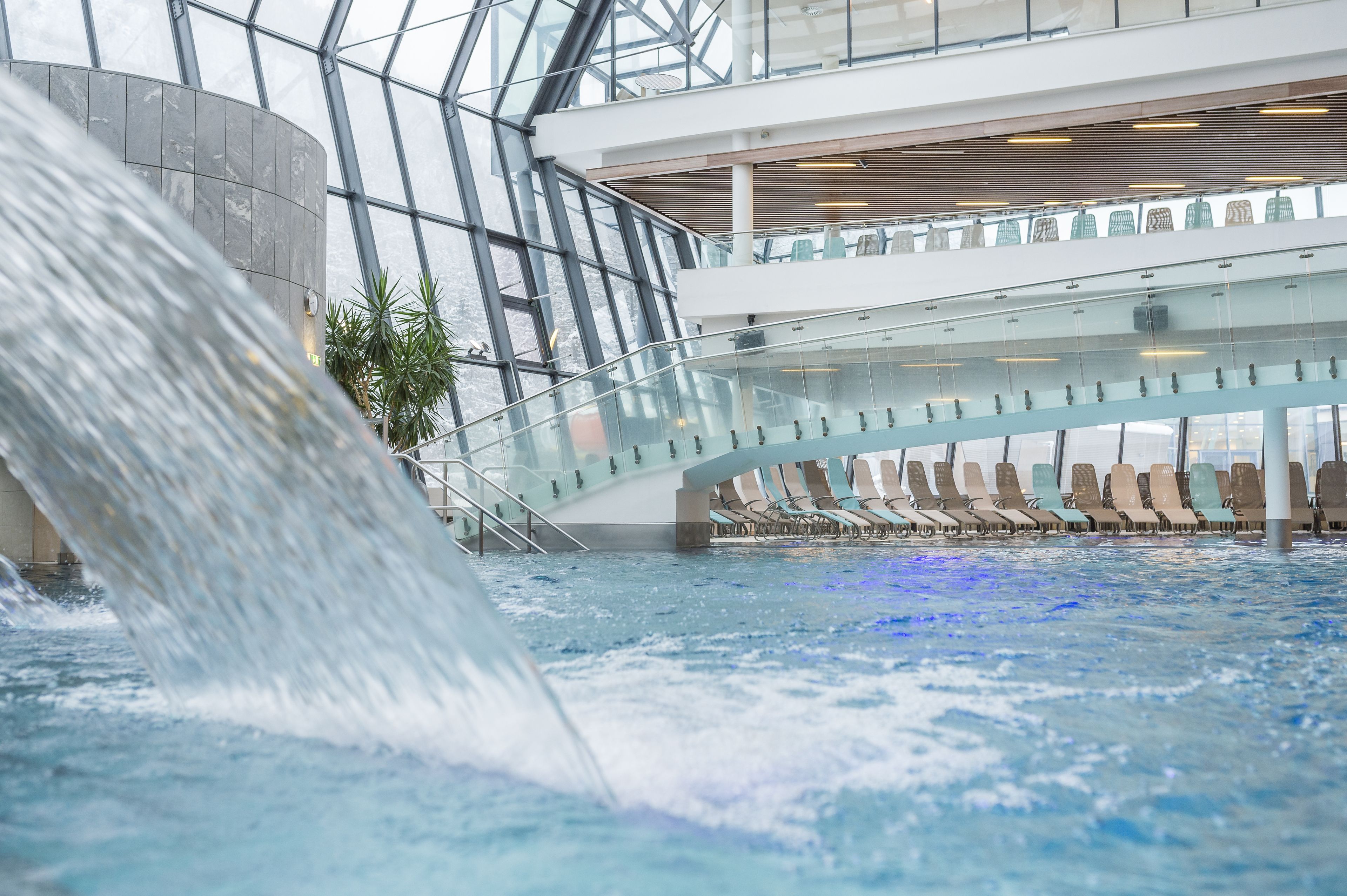 Una piscina interna moderna con cascata, ringhiere in vetro e zone relax.