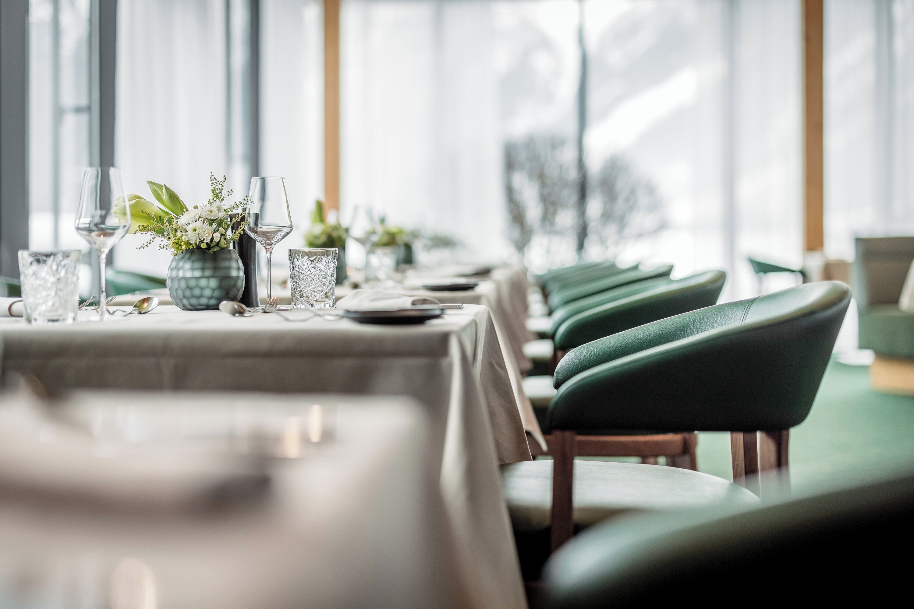 A restaurant with tables set for dining, featuring green upholstered chairs and a vase with flowers.