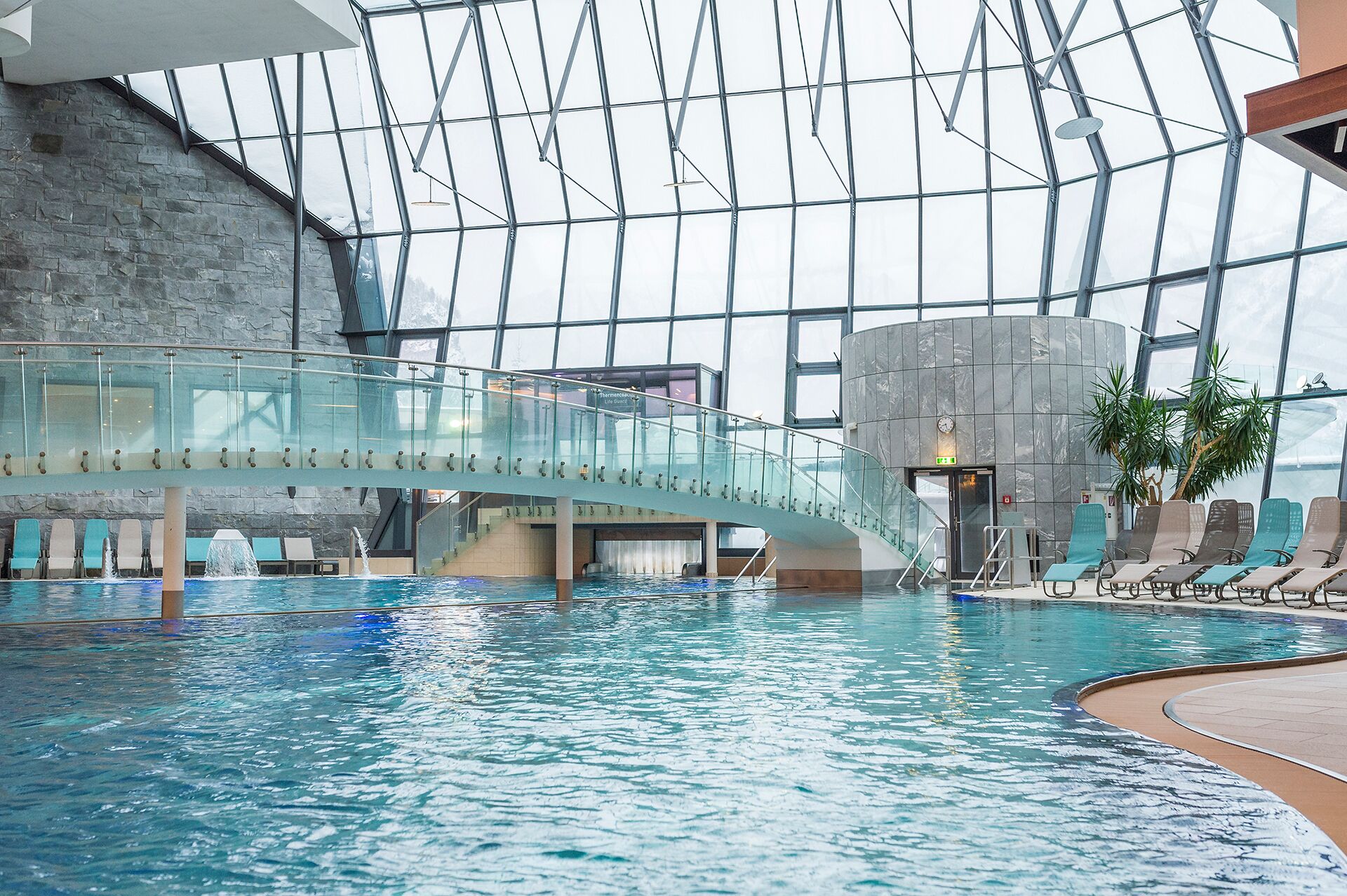 Una piscina interior con un puente de cristal, sillas de descanso y una pared de piedra.