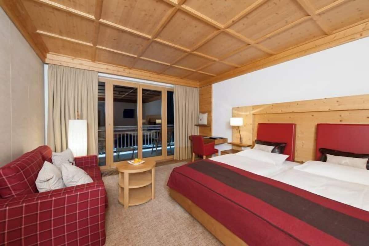 A hotel room with a wooden ceiling, a large bed with red headboards, a red plaid sofa, a small desk, and a view through a glass door.