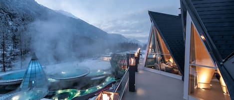 A modern hotel with a glass-enclosed infinity pool, a hot spring area, and a view of snow-covered mountains.