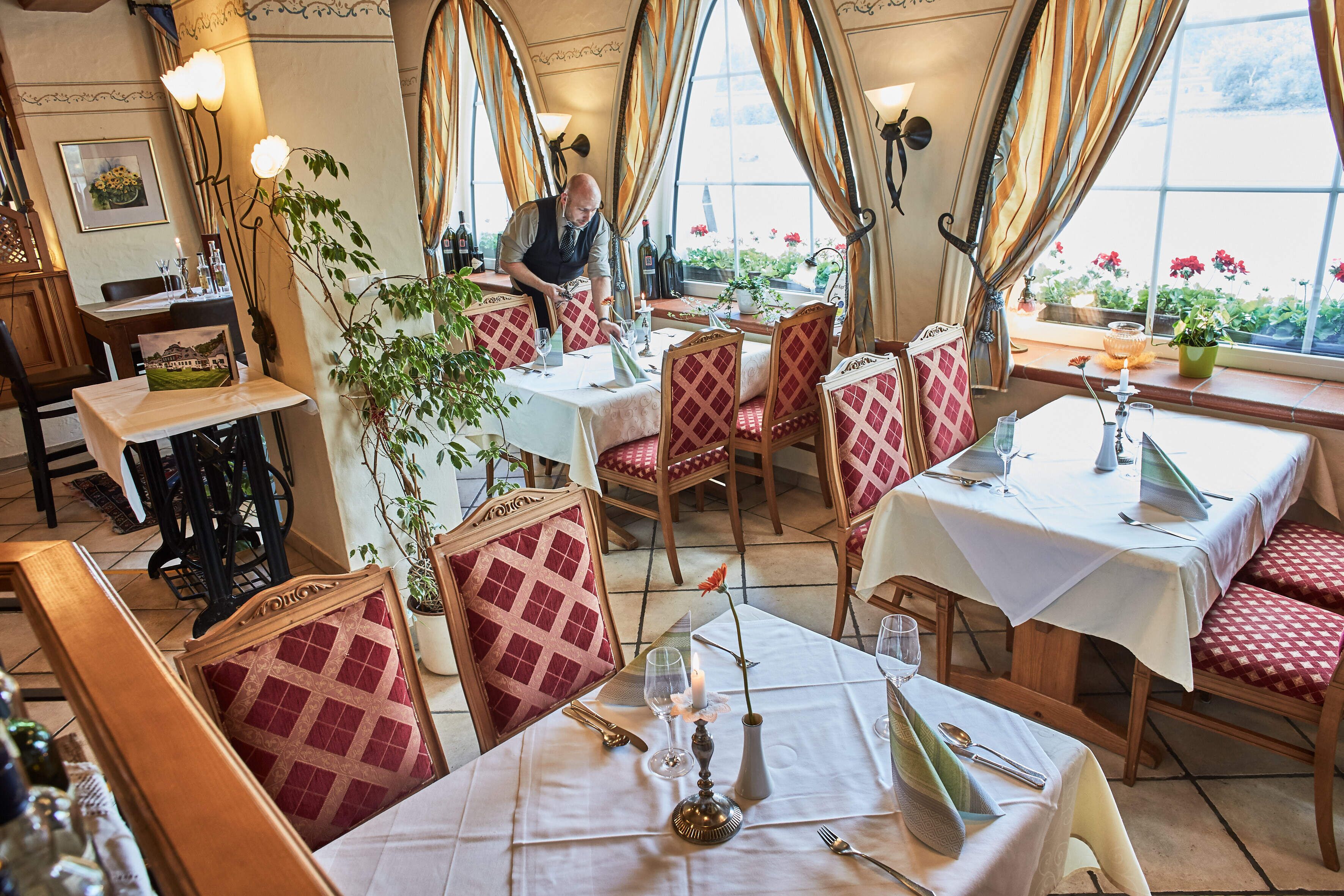 A restaurant with red and white checkered chairs, tables set with wine glasses, and a waiter attending to guests.